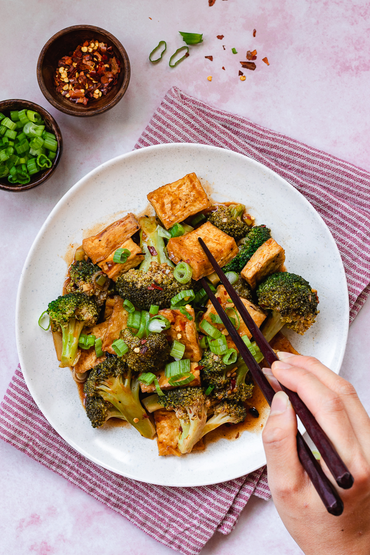 chili sauce tofu and broccoli served on a plate, hand holding chopsticks reaching for a piece of tofu