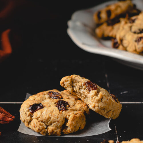 two oatmeal cookies staggered on each other, plate of cookies in background