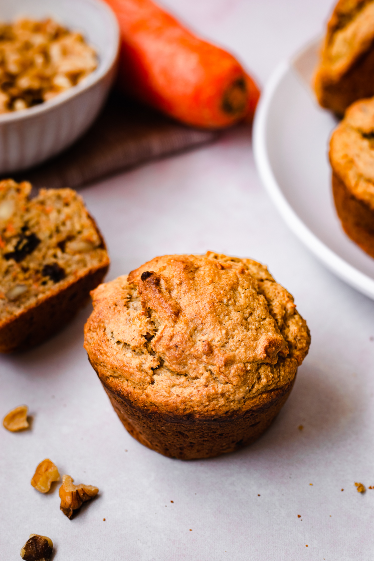 a vegan carrot banana muffin surrounded by muffins, walnuts, and carrots in the background