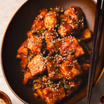 sweet and sour tofu in a bowl with chopsticks, pinch bowl of sesame seeds in the corner