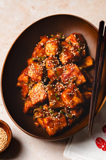 sweet and sour tofu in a bowl with chopsticks, pinch bowl of sesame seeds in the corner
