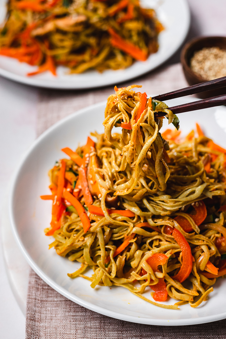 chopsticks holding a bite of edamame noodle stir fry above plate