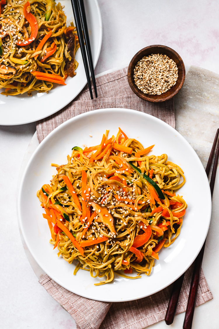 two plates of edamame noodle stir fry with a small bowl of sesame seeds