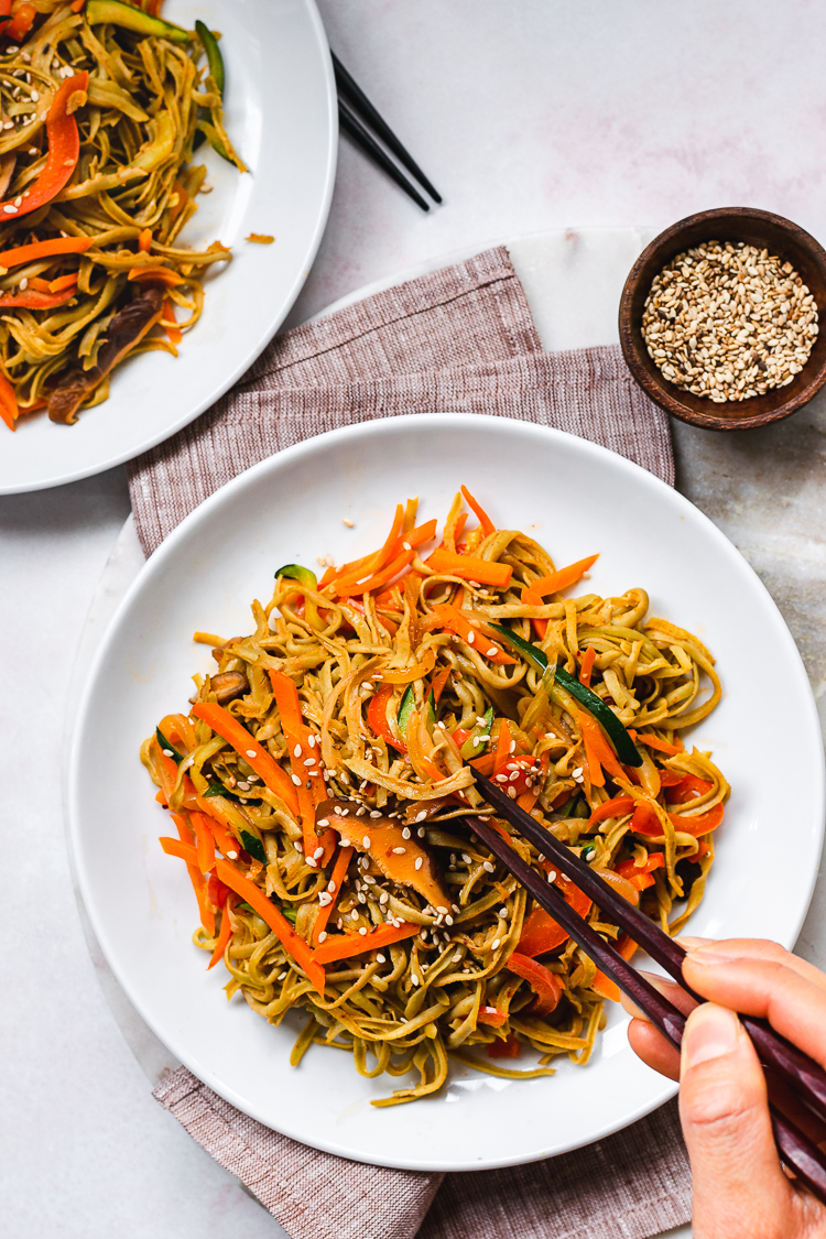 two plates of edamame noodle stir fry with hands on chopsticks reaching for the noodles