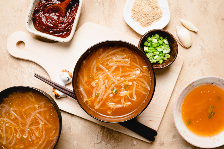 2 bowls of bean sprout miso soup with green onions, sesame seeds, and gochujang in different bowls around the soup