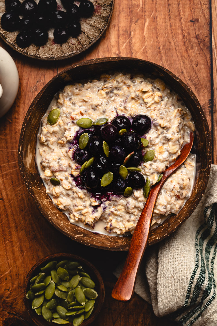 overnight oats in a bowl seen from overhead, topped with frozen blueberries and pumpkin seeds, spoon in bowl